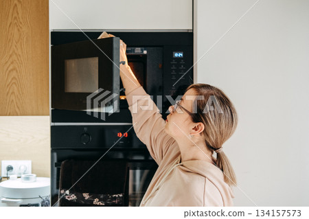 A woman wearing glasses and a hoodie reaches into a microwave oven built into a modern kitchen cabinet. Sustainable and Eco-Conscious Cleaning Alternatives, reducing paper waste, reusable cloths. A woman wearing glasses and a hoodie reaches into a microwave oven built into a modern kitchen cabinet. Sustainable and Eco-Conscious Cleaning Alternatives, reducing paper waste, reusable cloths. 134157573