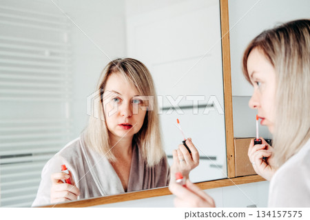 Woman applies lip gloss looking intently at her reflection in bathroom mirror. No-filter beauty, everyday makeup routine, realistic portrayal, morning getting ready. Woman applies lip gloss looking intently at her reflection in bathroom mirror. No-filter beauty, everyday makeup routine, realistic portrayal, morning getting ready. 134157575