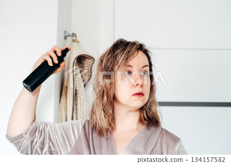 A woman in a robe sprays a product onto her damp shoulder length hair, looking into the distance with a serious expression. Hair loss concern, hair maintenance, clean haircare, self appraisal. 134157582