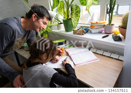 A schoolgirl girl studies with a tutor at home, additional lesson preparation, dad helps his daughter with lessons A schoolgirl girl studies with a tutor at home, additional lesson preparation, dad helps his daughter with lessons 134157811