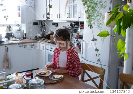 The girl has breakfast in the kitchen with scrambled eggs and a sandwich at the table in the morning The girl has breakfast in the kitchen with scrambled eggs and a sandwich at the table in the morning 134157843