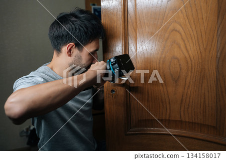 Close-up of professional handyman installing new door lock into wooden door frame with electric screwdriver, focusing on task of home repair and improvement. Concept of domestic tasks. 134158017