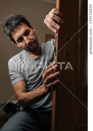 Man installing new door lock mechanism in modern home, providing security and ensuring proper home maintenance during repair and renovation work. Man installing new door lock mechanism in modern home, providing security and ensuring proper home maintenance during repair and renovation work. 134158026