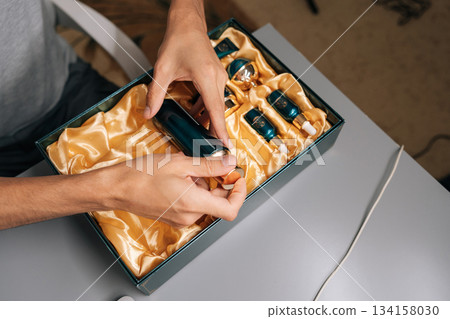 Close-up top view of man hands opening bottle from dark green gift box, revealing elegant collection of male grooming and skin care products nestled in golden silk inside. Close-up top view of man hands opening bottle from dark green gift box, revealing elegant collection of male grooming and skin care products nestled in golden silk inside. 134158030