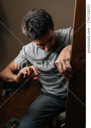 Vertical shot of professional technician male fixing door lock with screwdriver, performing installation and repair work on mechanism to ensure security and functionality within house. Vertical shot of professional technician male fixing door lock with screwdriver, performing installation and repair work on mechanism to ensure security and functionality within house. 134158031
