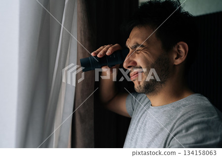 Bearded young man peers through white curtains holding monocular, intently watching street outside. Close-up of domestic scene suggesting secrecy, surveillance, investigation, curiosity and suspicion. 134158063