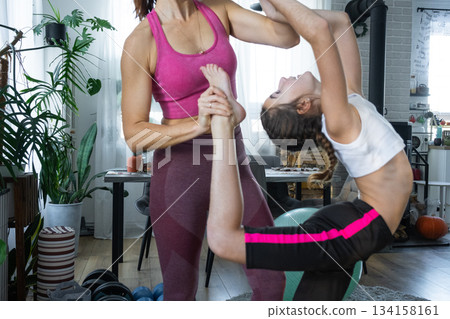 The girl does gymnastic stretching with the help of a coach at home, a home tutor, a mother trains her daughter 134158161