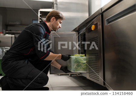 Male cook places large container of lettuce placed into refrigerator Male cook places large container of lettuce placed into refrigerator 134158460