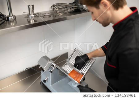 Man in uniform slicing meat with professional slicer Man in uniform slicing meat with professional slicer 134158461