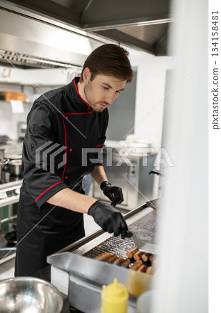 Cook preparing sausages on hot stainless steel surface in kitchen 134158481