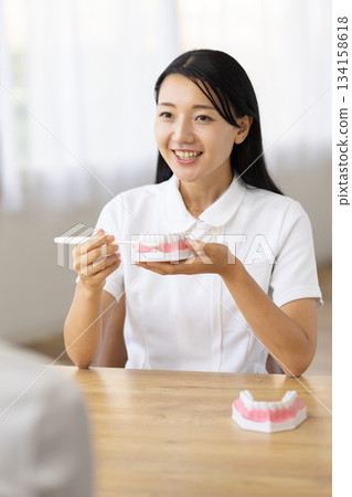 一位身穿白大褂的女士面帶微笑，一邊交談，一邊拿著牙齒模型和牙刷。 134158618