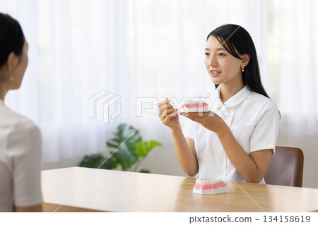 A woman in a white coat smiling and talking while holding a tooth model and a toothbrush 134158619