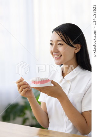 A woman in a white coat smiling and talking while holding a tooth model and a toothbrush 134158622