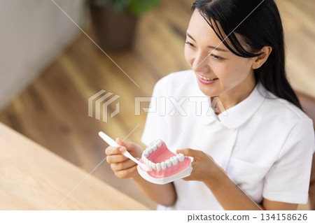 A woman in a white coat smiling and talking while holding a tooth model and a toothbrush A woman in a white coat smiling and talking while holding a tooth model and a toothbrush 134158626