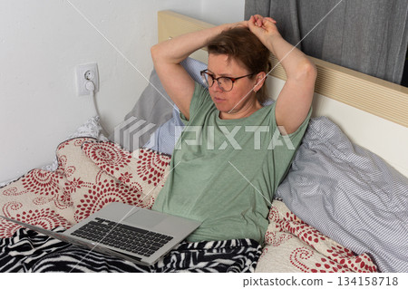 A woman sits on her bed in a bedroom using a laptop. She looks focused as she works and holds a cup in one hand. The setting shows an ordinary morning routine A woman sits on her bed in a bedroom using a laptop. She looks focused as she works and holds a cup in one hand. The setting shows an ordinary morning routine 134158718