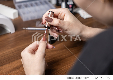 Woman's hand applying natural brown gel nail polish to her fingernail for a self-manicure, promoting personal beauty and self-care Woman's hand applying natural brown gel nail polish to her fingernail for a self-manicure, promoting personal beauty and self-care 134159420