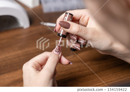 A woman applies brown gel nail polish to her own long nails during a home manicure session, emphasizing personal care and beauty routine 134159422