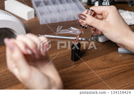 Woman's hand applies brown gel nail polish from bottle, surrounded by manicure tools, UV lamp, buffer, for home beauty self-care Woman's hand applies brown gel nail polish from bottle, surrounded by manicure tools, UV lamp, buffer, for home beauty self-care 134159424