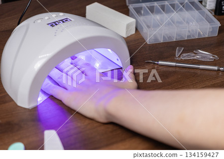 Woman's hand curing glitter purple gel nails in a modern LED UV lamp, performing a personal self-manicure beauty treatment Woman's hand curing glitter purple gel nails in a modern LED UV lamp, performing a personal self-manicure beauty treatment 134159427