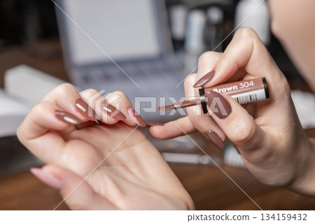 Woman's hands performing a meticulous personal manicure, carefully applying Brown gel polish on nail extension upper forms, emphasizing DIY beauty 134159432