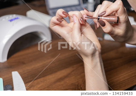 Woman applying gel nail polish to her extended nails by herself with Brown 304 color, doing a home manicure for personal beauty care 134159433