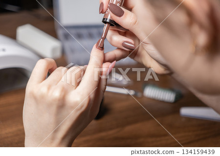 Woman's hand applying brown gel polish to a fingernail, creating a chic do-it-yourself manicure, symbolizing personal beauty, self-care, and relaxing at-home pampering Woman's hand applying brown gel polish to a fingernail, creating a chic do-it-yourself manicure, symbolizing personal beauty, self-care, and relaxing at-home pampering 134159439