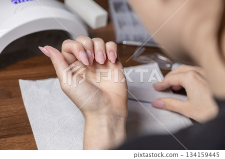 Woman doing her own manicure at home with an unfinished gel nail application, showcasing personal beauty care and a DIY nail treatment process 134159445