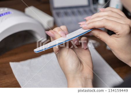 Woman's hands carefully filing her elegant nude gel nails with a nail file at home, performing a precise DIY manicure for personal beauty and self-grooming Woman's hands carefully filing her elegant nude gel nails with a nail file at home, performing a precise DIY manicure for personal beauty and self-grooming 134159449