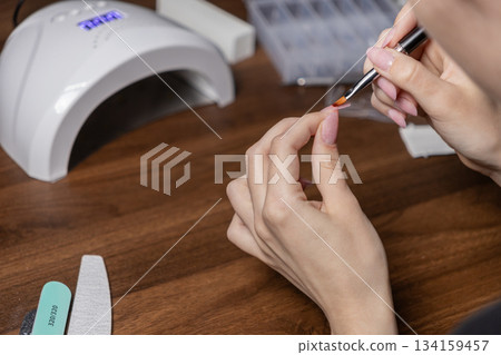 Female hand applying gel polish to fingernail during a self-care manicure, utilizing a UV lamp and professional nail art tools on wooden desk 134159457