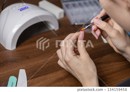 Woman applying polygel onto a nail tip for a DIY manicure by oneself at home, utilizing a UV lamp for a beauty routine 134159458