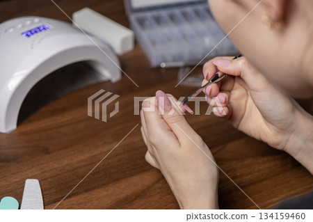 Woman's hands applying gel nail extension by oneself using a tool, performing a DIY home manicure with a UV lamp for personal beauty 134159460