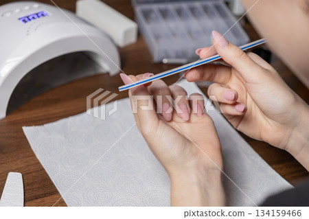 Young woman manicuring by herself, meticulously filing her pink gel nails with a detailed file at home, surrounded by professional beauty tools for self-care Young woman manicuring by herself, meticulously filing her pink gel nails with a detailed file at home, surrounded by professional beauty tools for self-care 134159466