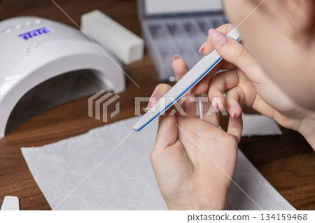 Woman diligently filing nails with a manicure file, performing a professional-grade DIY gel polish application by oneself at home for beauty and self-care Woman diligently filing nails with a manicure file, performing a professional-grade DIY gel polish application by oneself at home for beauty and self-care 134159468