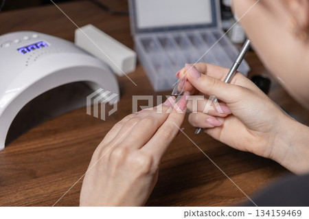 Adult woman performs self-manicure, carefully applying a clear nail extension upper form with pink gel, utilizing a modern UV LED lamp and essential beauty tools Adult woman performs self-manicure, carefully applying a clear nail extension upper form with pink gel, utilizing a modern UV LED lamp and essential beauty tools 134159469
