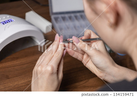Woman's hands applying gel to a nail extension upper form, performing a detailed self-manicure with professional home beauty tools and a nail lamp Woman's hands applying gel to a nail extension upper form, performing a detailed self-manicure with professional home beauty tools and a nail lamp 134159471