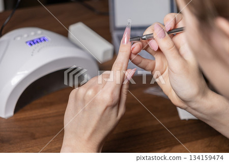 Young woman carefully performing a DIY nail extension manicure on herself, applying an upper form with a tool for perfect, elegant long nails Young woman carefully performing a DIY nail extension manicure on herself, applying an upper form with a tool for perfect, elegant long nails 134159474