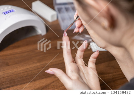 Woman meticulously applying artificial gel nail extension tip with tweezers to her finger during self-care home manicure, using a professional UV lamp Woman meticulously applying artificial gel nail extension tip with tweezers to her finger during self-care home manicure, using a professional UV lamp 134159482