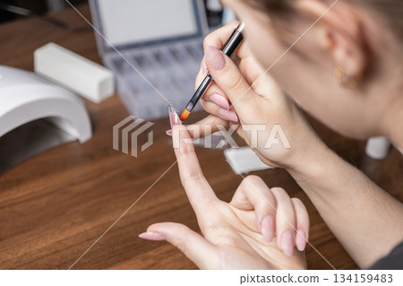 Woman carefully applying gel with a small brush onto a clear artificial nail tip for a modern DIY manicure, embodying personal self-care and home beauty 134159483