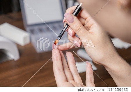 Woman performing self-manicure, applying clear gel or decorative element to a long artificial nude nail with a brush 134159485