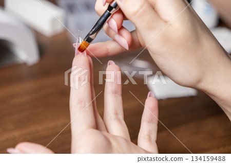 Woman's hands meticulously applying clear gel with a brush to an artificial nail extension tip, demonstrating a DIY manicure process for a perfect self-care beauty routine Woman's hands meticulously applying clear gel with a brush to an artificial nail extension tip, demonstrating a DIY manicure process for a perfect self-care beauty routine 134159488