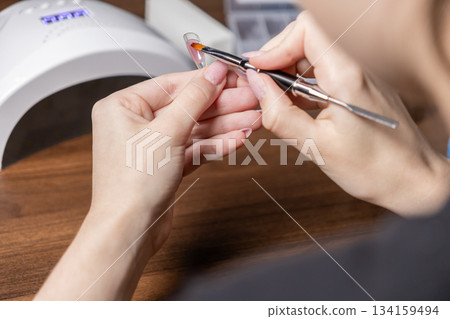 Woman's hands applying pink builder gel with a professional brush onto a clear nail extension upper form, ideal for a DIY home manicure Woman's hands applying pink builder gel with a professional brush onto a clear nail extension upper form, ideal for a DIY home manicure 134159494