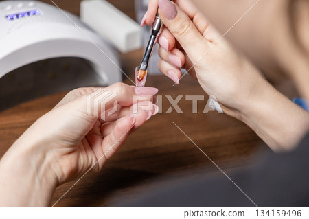 Woman's hands applying clear nail gel polish with a brush onto an upper form for self-manicure extension, creating a long-lasting beauty treatment at home 134159496