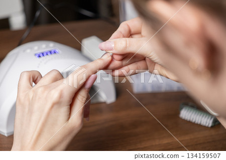 Woman skillfully applies her own gel nail extension using modern upper forms and a professional UV lamp, perfecting a DIY manicure for self-care beauty 134159507
