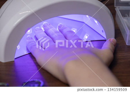 A female hand with a stylish French manicure curing under a purple UV LED nail lamp, representing personal beauty care and DIY home salon treatments 134159509