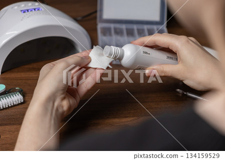 Woman's hands pouring 100ml liquid from a bottle onto a cotton pad, performing a detailed self-manicure for personal nail care and home beauty 134159529