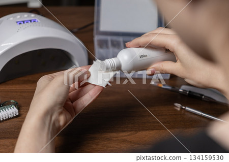 Hands pouring nail preparation liquid from a white 100ml bottle onto a cotton pad, performing a detailed gel manicure by oneself at home 134159530