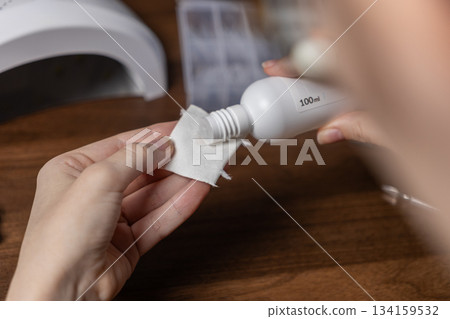 A woman's hands meticulously prepare for a home gel nail manicure by oneself, carefully pouring cleansing solution from a white bottle onto a cotton pad A woman's hands meticulously prepare for a home gel nail manicure by oneself, carefully pouring cleansing solution from a white bottle onto a cotton pad 134159532