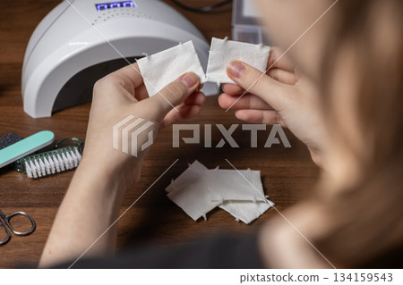 Woman's hands preparing cotton pads for self-manicure, surrounded by essential nail care tools, including a modern UV lamp Woman's hands preparing cotton pads for self-manicure, surrounded by essential nail care tools, including a modern UV lamp 134159543