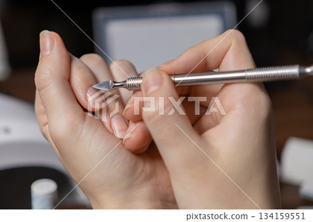 Close-up of a person performing a meticulous personal manicure by oneself, using a metal cuticle pusher tool for precise natural nail care, promoting hygiene and a wellness beauty routine 134159551