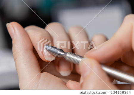 A womans hand skillfully performs a manicure by oneself, pushing back the natural cuticle with a metal tool for effective personal nail care 134159554
